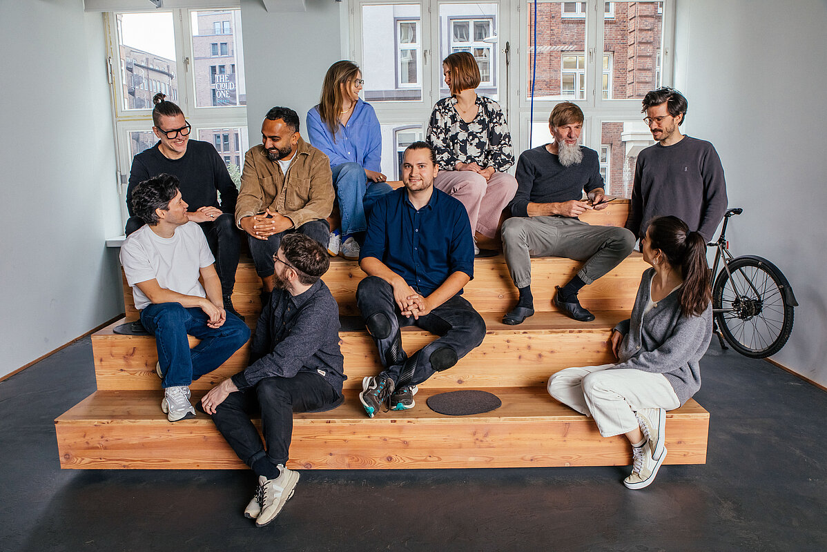 Eine Gruppe von neun Erwachsenen, die auf einer breiten Holztreppe oder gestuften Sitzgelegenheit in einem modernen Innenraum posiert. Die Gruppe ist locker angeordnet, einige Personen unterhalten sich, während andere in die Kamera schauen. Die Kleidung ist lässig-elegant. Im Hintergrund ist eine große Fensterfront mit Blick auf Backsteingebäude zu sehen. Rechts neben der Gruppe steht ein Fahrrad.