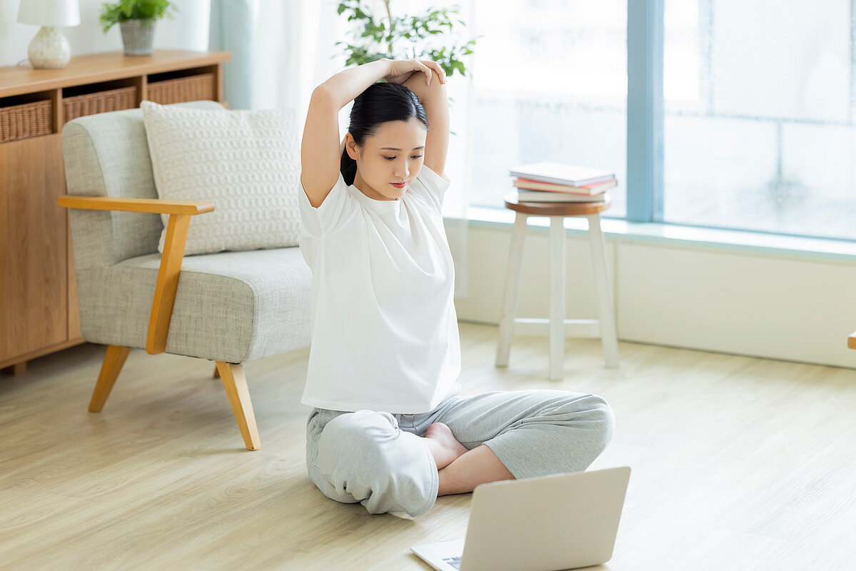 Eine Frau sitzt im Schneidersitz auf einem Holzboden in einem hellen Wohnzimmer. Sie trägt ein weißes T-Shirt und graue Jogginghosen und macht eine Dehnübung, indem sie ihre Arme über den Kopf streckt und die Hände zusammenführt. Vor ihr liegt ein geöffneter Laptop, was darauf hindeutet, dass sie einem Online-Workout oder Yoga-Kurs folgt. Im Hintergrund sind ein beigefarbener Sessel, ein Holzschrank und ein großes Fenster zu sehen, durch das helles Licht einfällt.