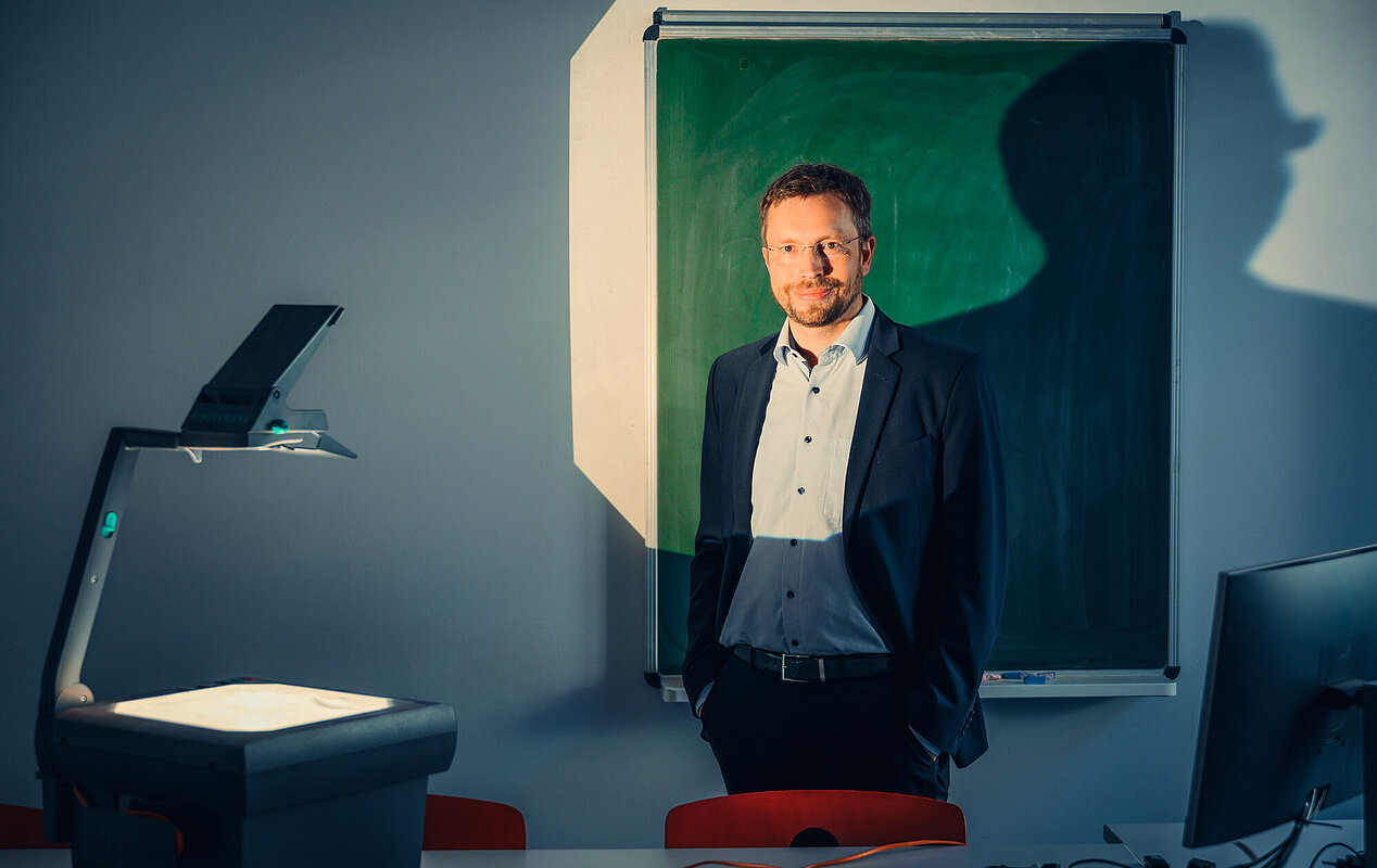 Ein Mann mittleren Alters steht in einem leicht abgedunkelten Raum, wahrscheinlich einem Klassenzimmer oder Vortragssaal. Er trägt ein dunkles Sakko über einem hellblauen oder grauen Hemd und blickt nach vorne. Rechts hinter ihm hängt eine grüne Kreidetafel, auf die ein dunkler Schatten fällt. Links neben ihm steht ein Dokumentenkamera-Projektor, dessen Licht auf den Schreibtisch leuchtet. Der Raum hat graue Wände.