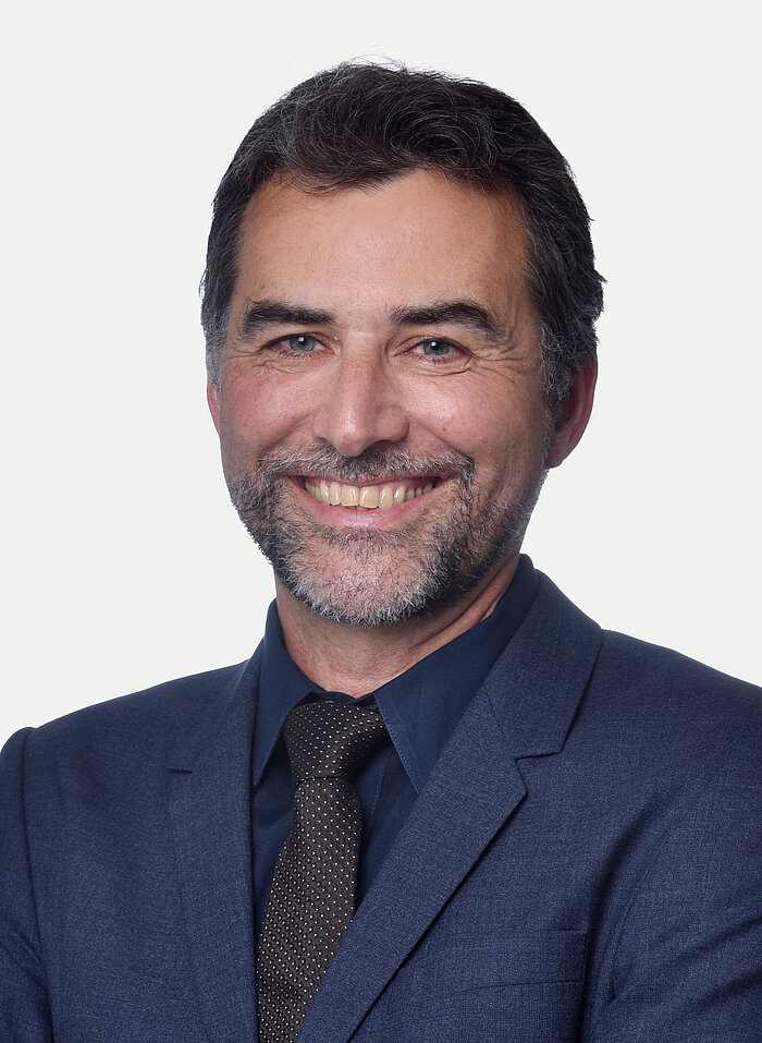 Porträtaufnahme eines lächelnden Mannes mittleren Alters mit dunklem Haar und Bart. Er trägt einen dunkelblauen Anzug, ein dunkles Hemd und eine dunkle Krawatte. Er blickt direkt in die Kamera. Der Hintergrund ist ein schlichter, heller Grauton.