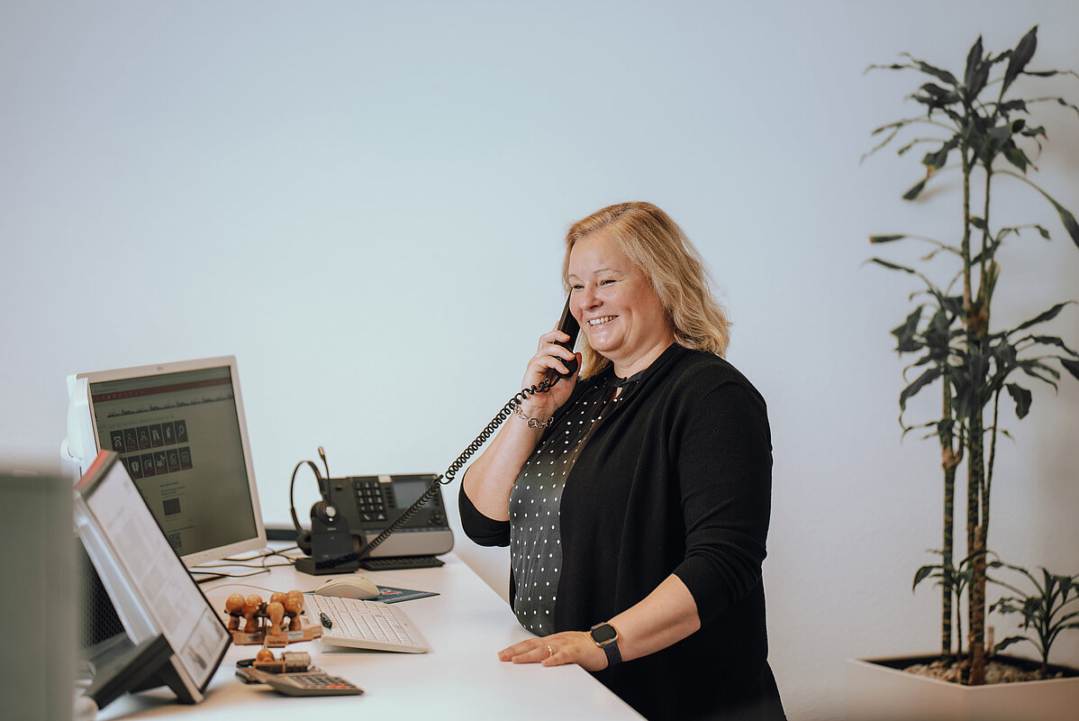 Eine Frau steht lächelnd an einem Schreibtisch und telefoniert. Vor ihr befinden sich ein Computerbildschirm, ein Telefon und einige Stempel. Im Hintergrund ist eine Pflanze zu sehen.