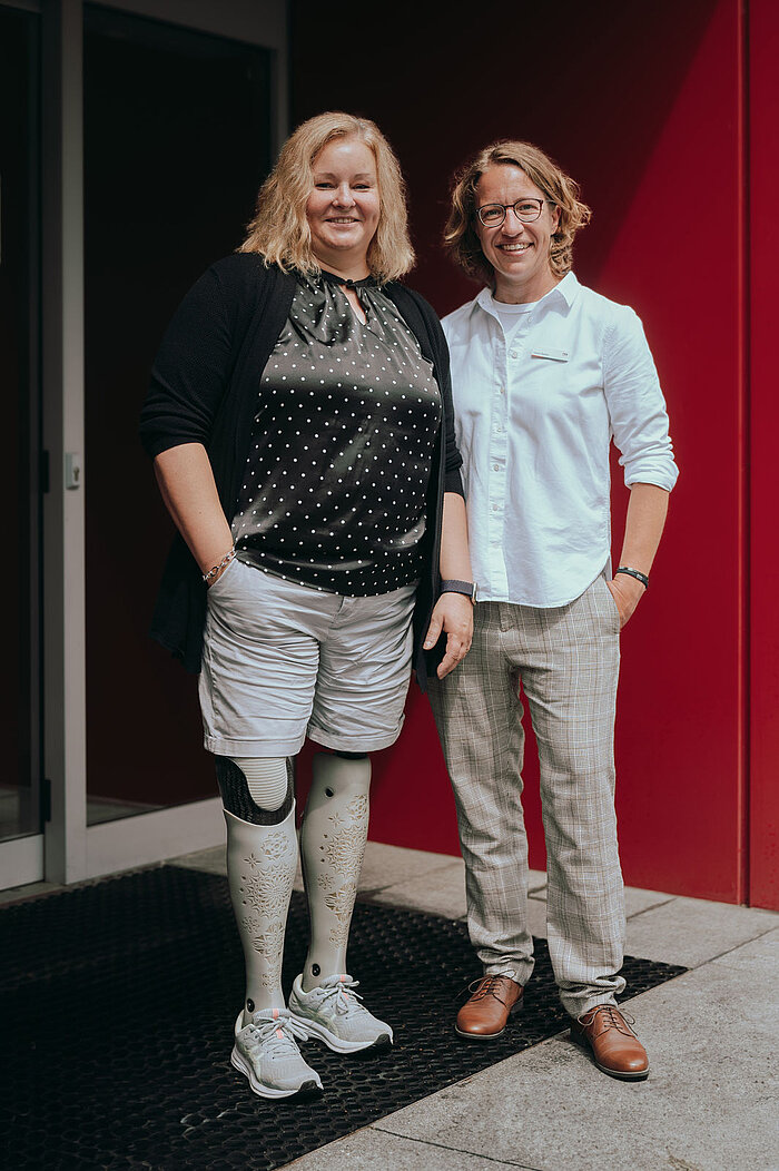 Zwei Frauen stehen nebeneinander im Stehen und lächeln in die Kamera. Die Frau links trägt einen schwarzen Cardigan über einem gemusterten Oberteil, helle Shorts und Sportschuhe. Sie trägt eine Beinprothese, die deutlich sichtbar ist. Die Frau rechts trägt ein weißes Hemd und helle Hosen. Beide stehen vor einem Eingangsbereich; links eine Glastür, rechts eine leuchtend rote Wand.
