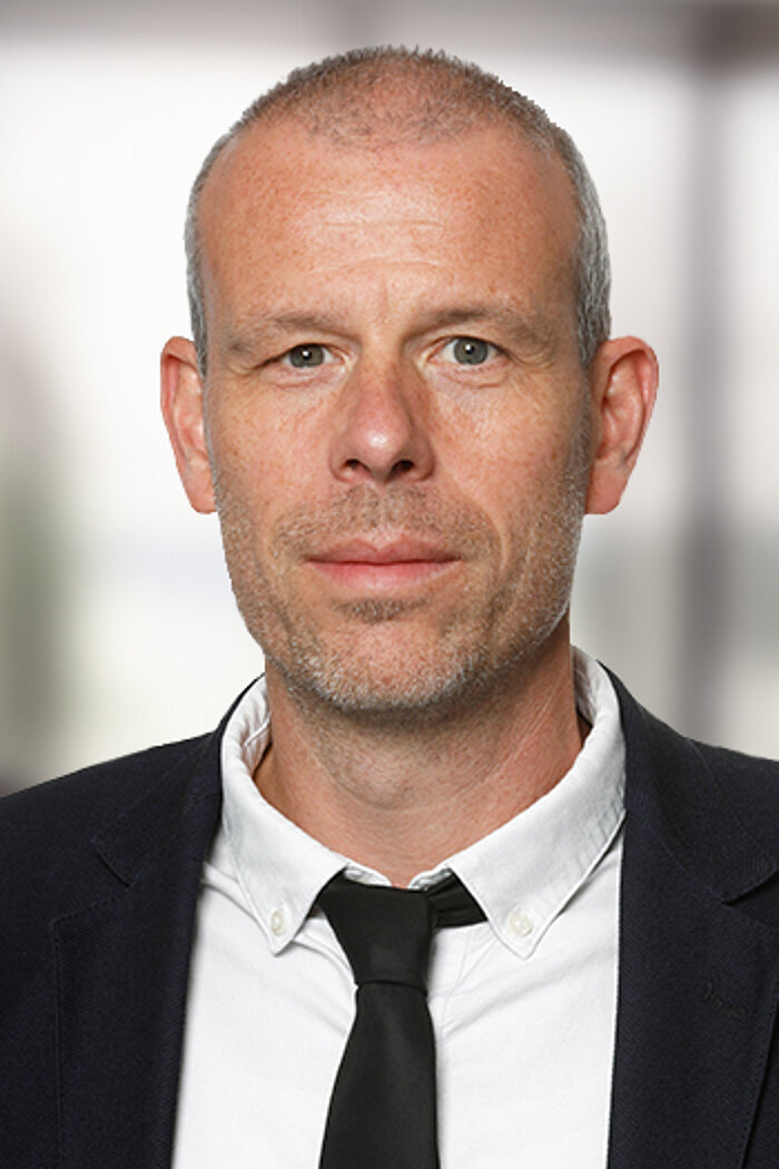 Foto: Portraitaufnahme von Herrn Dragano. Er schaut in die Kamera und lächelt leicht. Er trägt einen schwarzen Anzug, ein weißes Hemd und eine schwarze Kravatte.