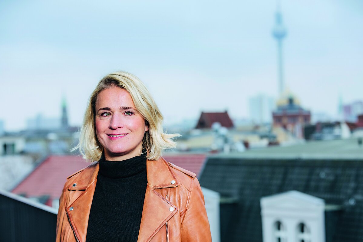 Eine Frau mit blonden Haaren und einer braunen Lederjacke steht lächelnd auf einer Dachterrasse. Im Hintergrund ist eine Stadtlandschaft mit einem Fernsehturm zu sehen.