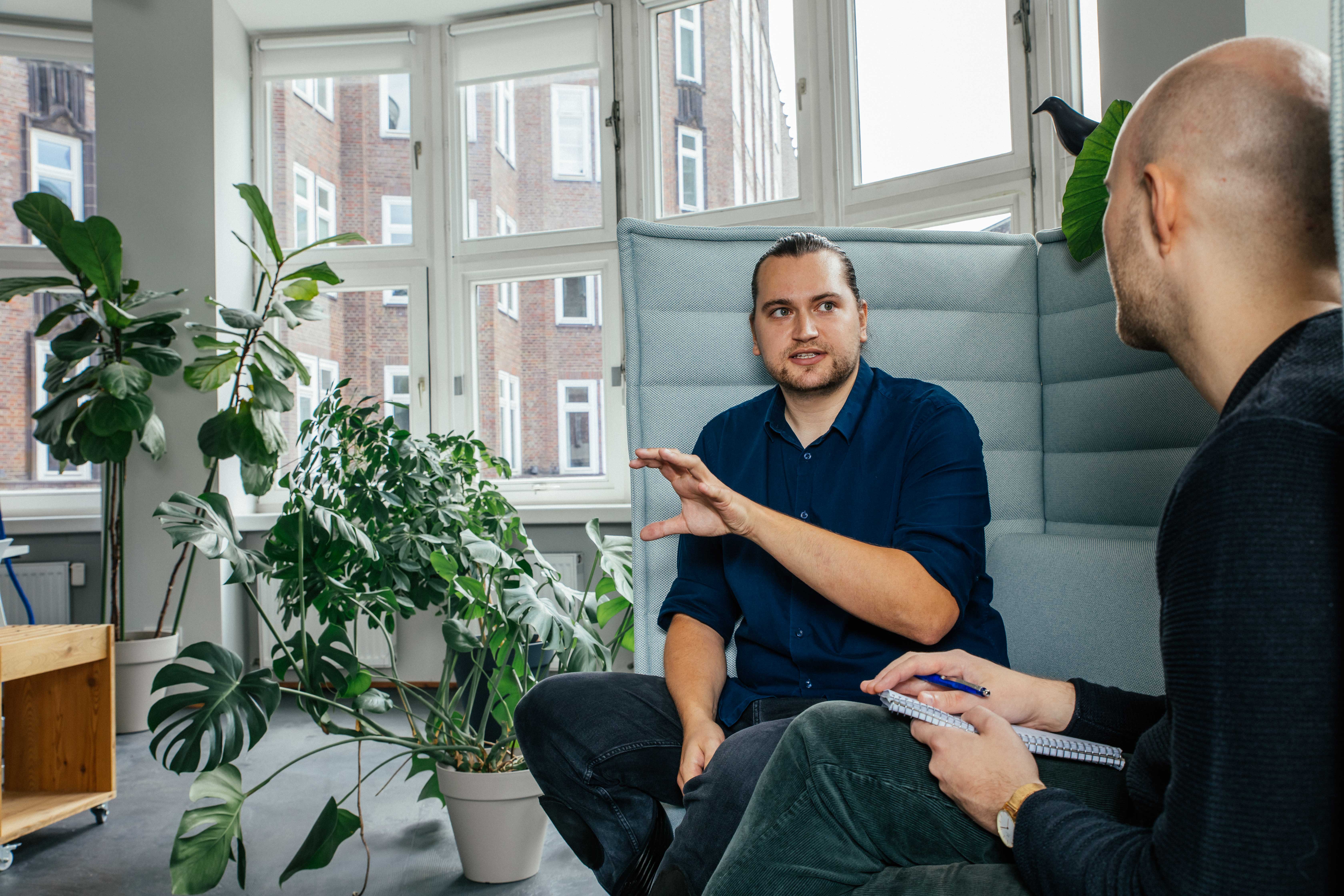 Zwei Männer sitzen in einem modernen Büro mit großen Fenstern und vielen Zimmerpflanzen und unterhalten sich. Der Mann in der Mitte des Bildes trägt ein dunkelblaues Hemd, sitzt auf einem gepolsterten, grauen Sitzmöbel und gestikuliert mit beiden Händen. Der zweite Mann sitzt ihm gegenüber, mit dem Rücken zur Kamera, und hält ein Notizbuch. Die Umgebung ist hell, mit großen Fenstern und verschiedenen Topfpflanzen wie Monstera und anderen Grünpflanzen.