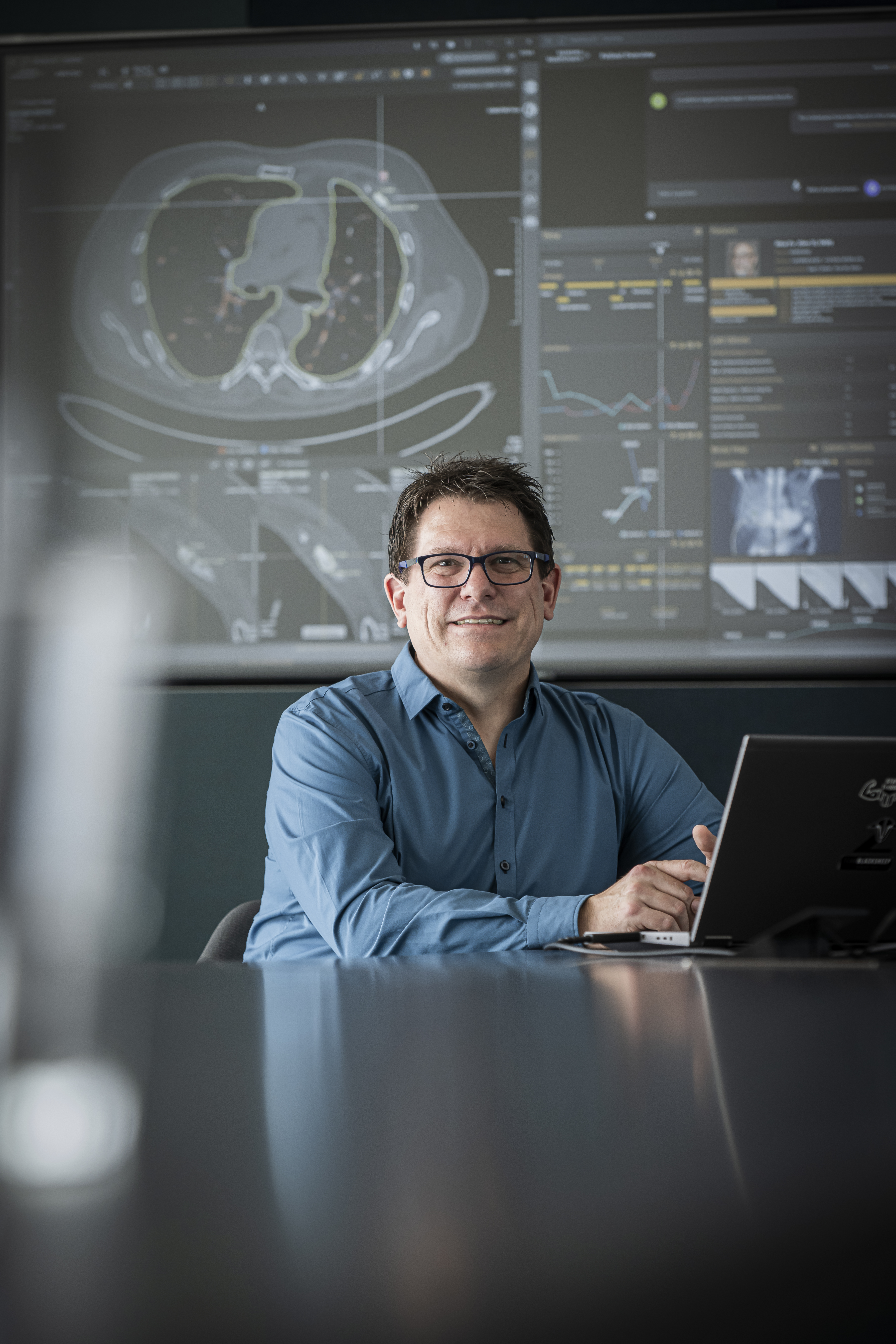 Potraitfoto von Herrn Thesen. Herr Thesen sitzt an einem Tisch und hat vor sich einen Laptop aufgeklappt. Er trägt ein blaues Hemd, hat kurze braune Haare und trägt eine Brille. Er lächelt in die Kamera. Im Hintergrund ist ein Whiteborad zu sehen auf dem ein KI-Tool von Siemens Healthineers zu sehen ist.