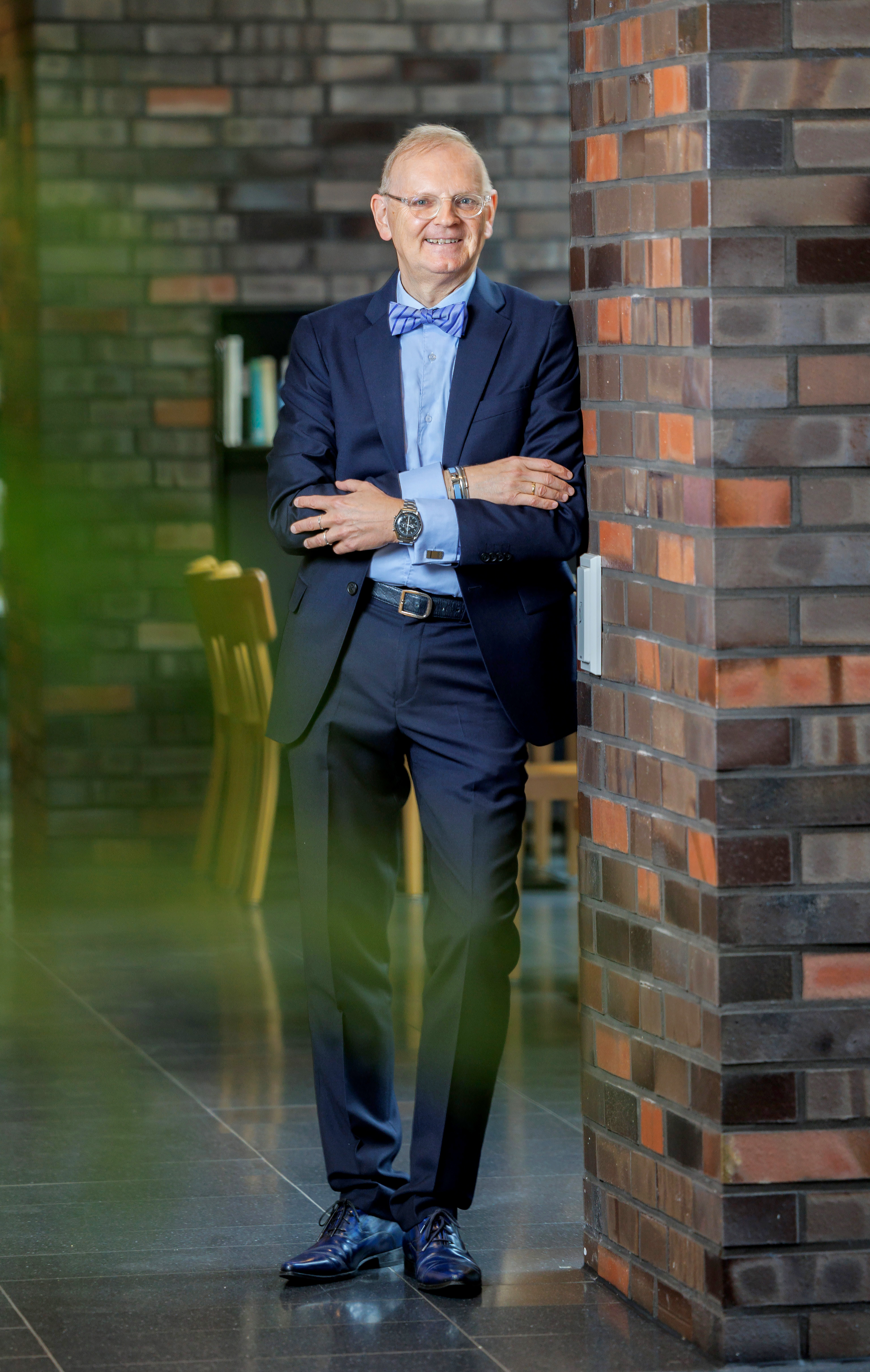 Portraitbild Herr Froese. Herr Froese steht an eine Backsteinmauer gelehnt. Seine Arme sind locker verschränkt. Er trägt einen dunkelblauen Anzug mit einem hellblauen Hemd und einer hellblauen Fliege. Er hat kurze weiße Haare und trägt eine Brille. Er lächelt in die Kamera.