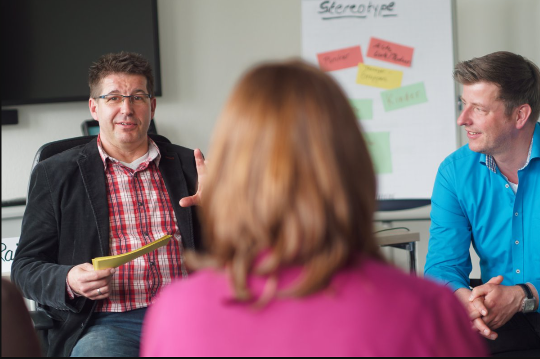 In einem Besprechungsraum sitzen drei Personen in einer Gesprächsrunde. Ein Mann in einem karierten Hemd und schwarzem Sakko spricht, während er ein Notizbuch hält. Eine Frau im Vordergrund hört zu. Ein weiterer Mann in einem blauen Hemd sitzt daneben und lächelt. Im Hintergrund ist ein Flipchart mit dem Titel "Stereotype" und farbigen Notizen zu sehen.