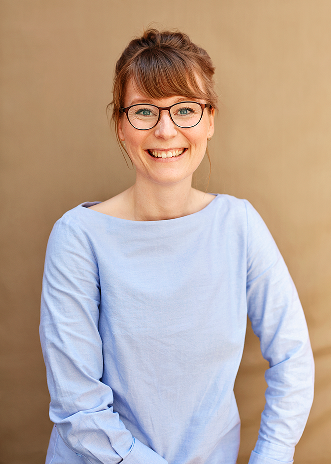Porträtaufnahme einer lächelnden Frau mit hochgestecktem Haar und Pony. Sie trägt eine Brille mit dunklem Rahmen und ein hellblaues Langarmshirt mit einem weiten, U-förmigen Ausschnitt. Der Hintergrund ist unscharf und hat eine neutrale, beigefarbene oder hellbraune Farbe.