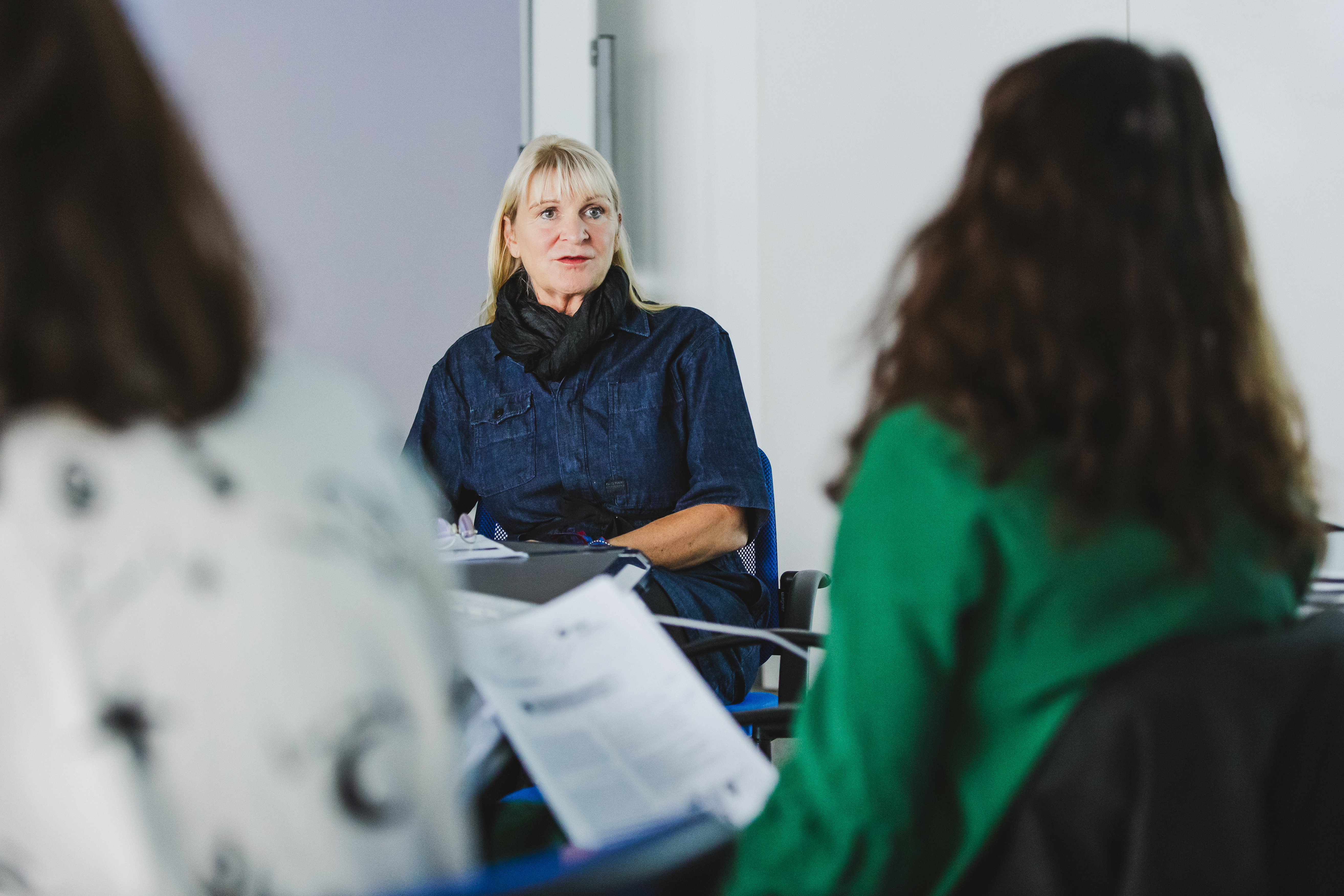 Eine blonde Frau mittleren Alters sitzt in einem hellen Besprechungsraum an einem Tisch und blickt nachdenklich in die Runde. Sie trägt ein dunkelblaues Denim-Hemd und einen schwarzen Schal. Sie ist umgeben von zwei anderen, unscharfen Personen, die mit dem Rücken zur Kamera sitzen. Die Frau im rechten Vordergrund hält Papiere in der Hand.
