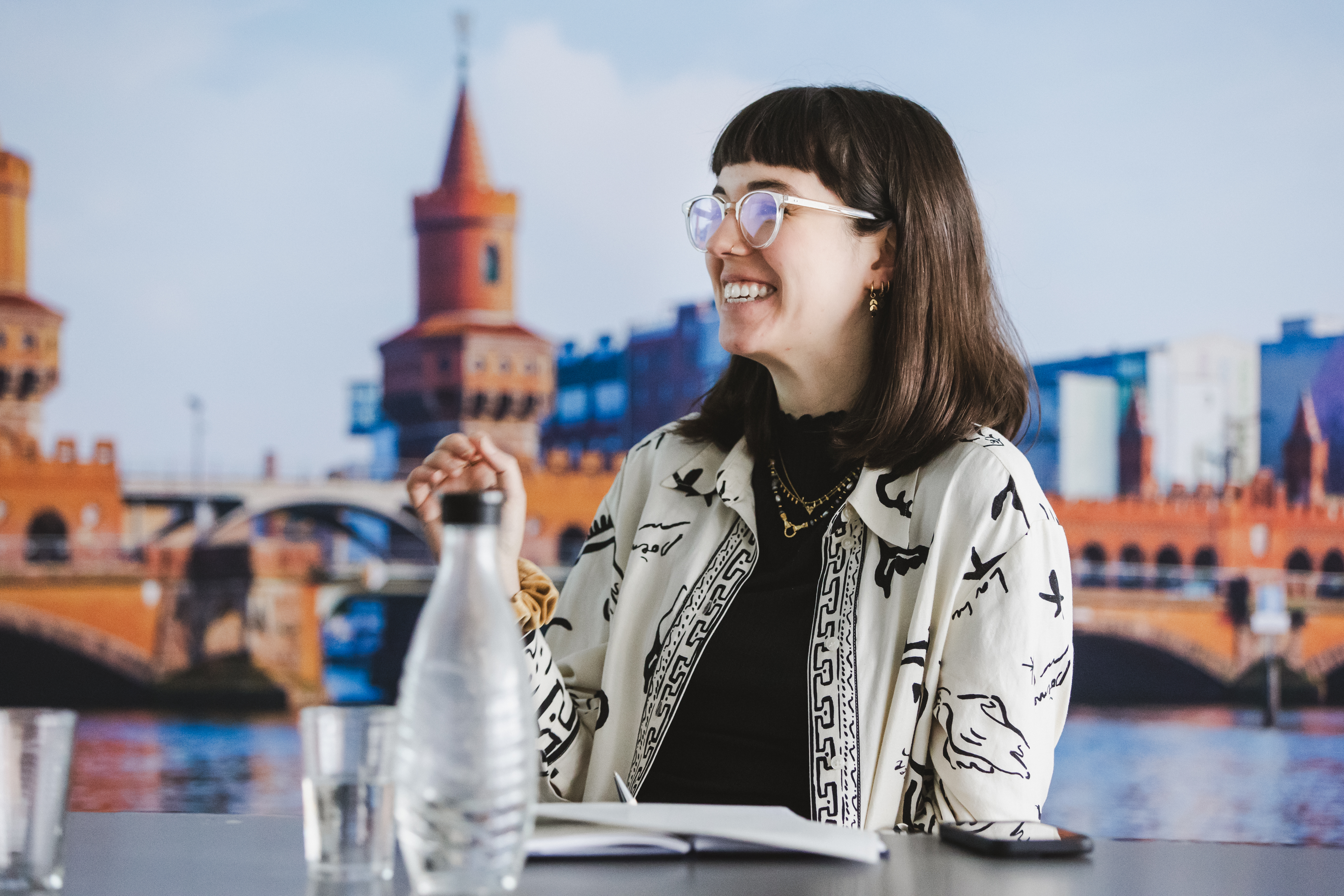 Eine junge Frau mit dunklem, schulterlangem Haar und Pony sitzt lächelnd und zur Seite blickend an einem Tisch. Sie trägt eine runde, helle Brille, einen schwarzen Rollkragenpullover und eine helle Jacke mit schwarzem, grafischem Muster. Vor ihr auf dem Tisch stehen Wassergläser und eine Wasserflasche. Im Hintergrund ist eine großflächige Darstellung der Oberbaumbrücke in Berlin zu sehen.