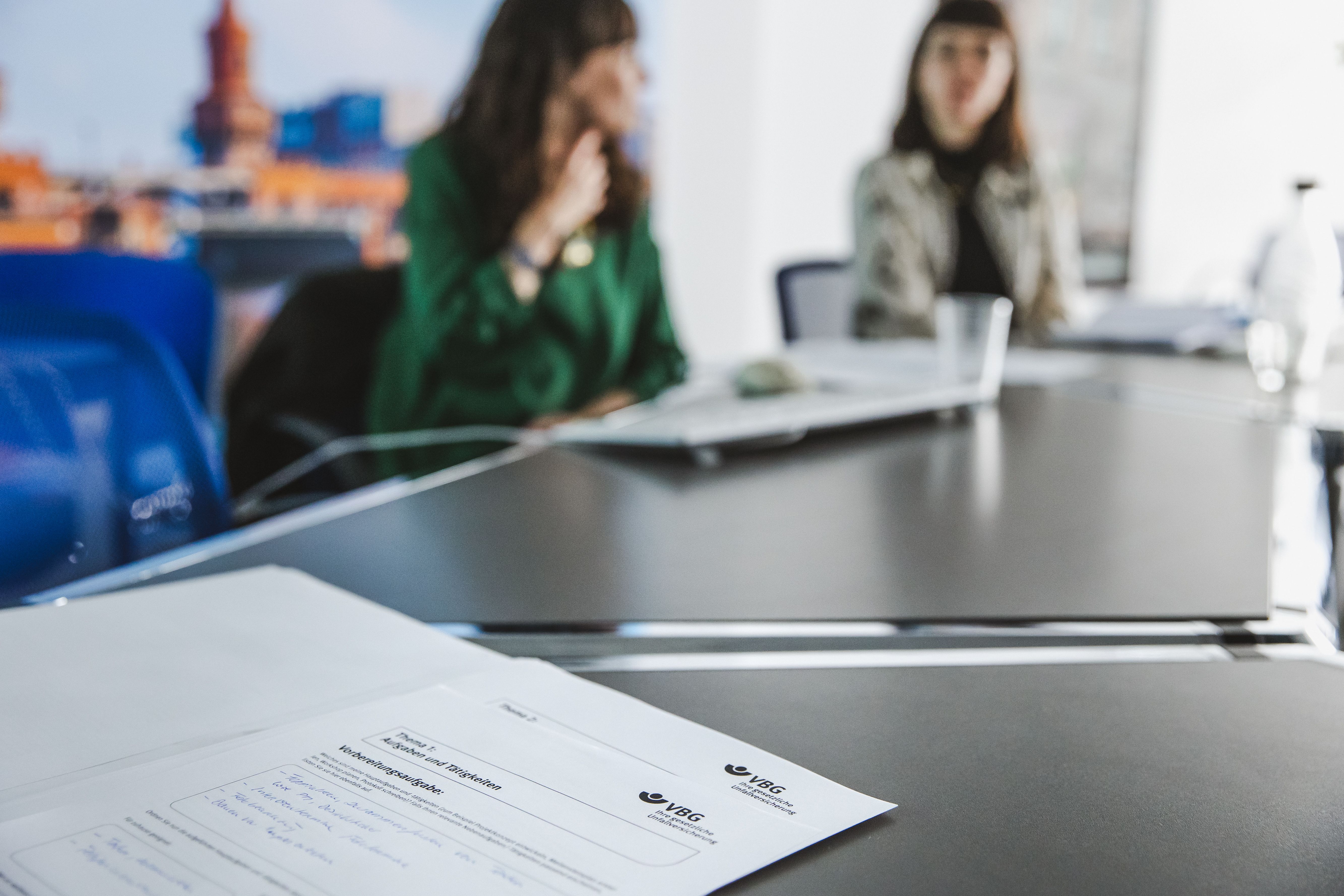 Nahaufnahme eines Dokumentes mit dem Logo der VBG und der Überschrift "Thema X: Aufgaben und Tätigkeiten" auf einem dunklen Besprechungstisch. Das Dokument enthält handschriftliche Notizen. Im unscharfen Hintergrund sitzen zwei Frauen an dem Tisch, die sich unterhalten. Eine der Frauen trägt ein grünes Oberteil, die andere ein hell gemustertes Oberteil. Der Hintergrund zeigt eine Wand mit einer unscharfen, großflächigen Stadtansicht.