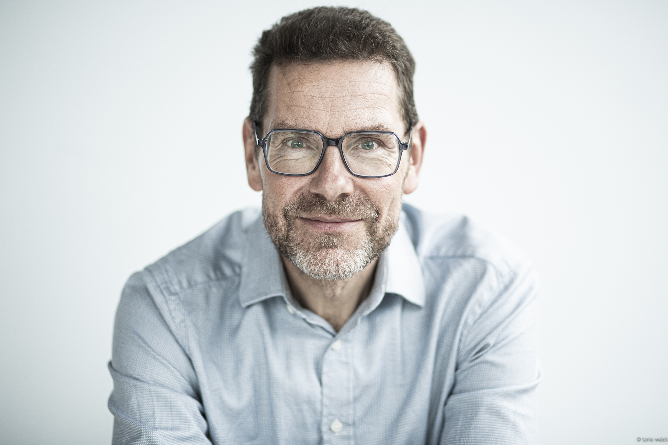Ein Mann mit Brille und grauem Hemd schaut freundlich in die Kamera. Der Hintergrund ist hell und unscharf.